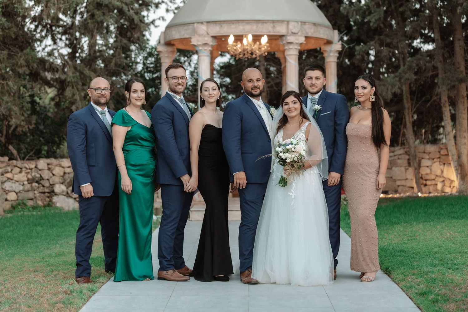 Wedding party poses together in formal attire on a manicured lawn with stone gazebo backdrop.