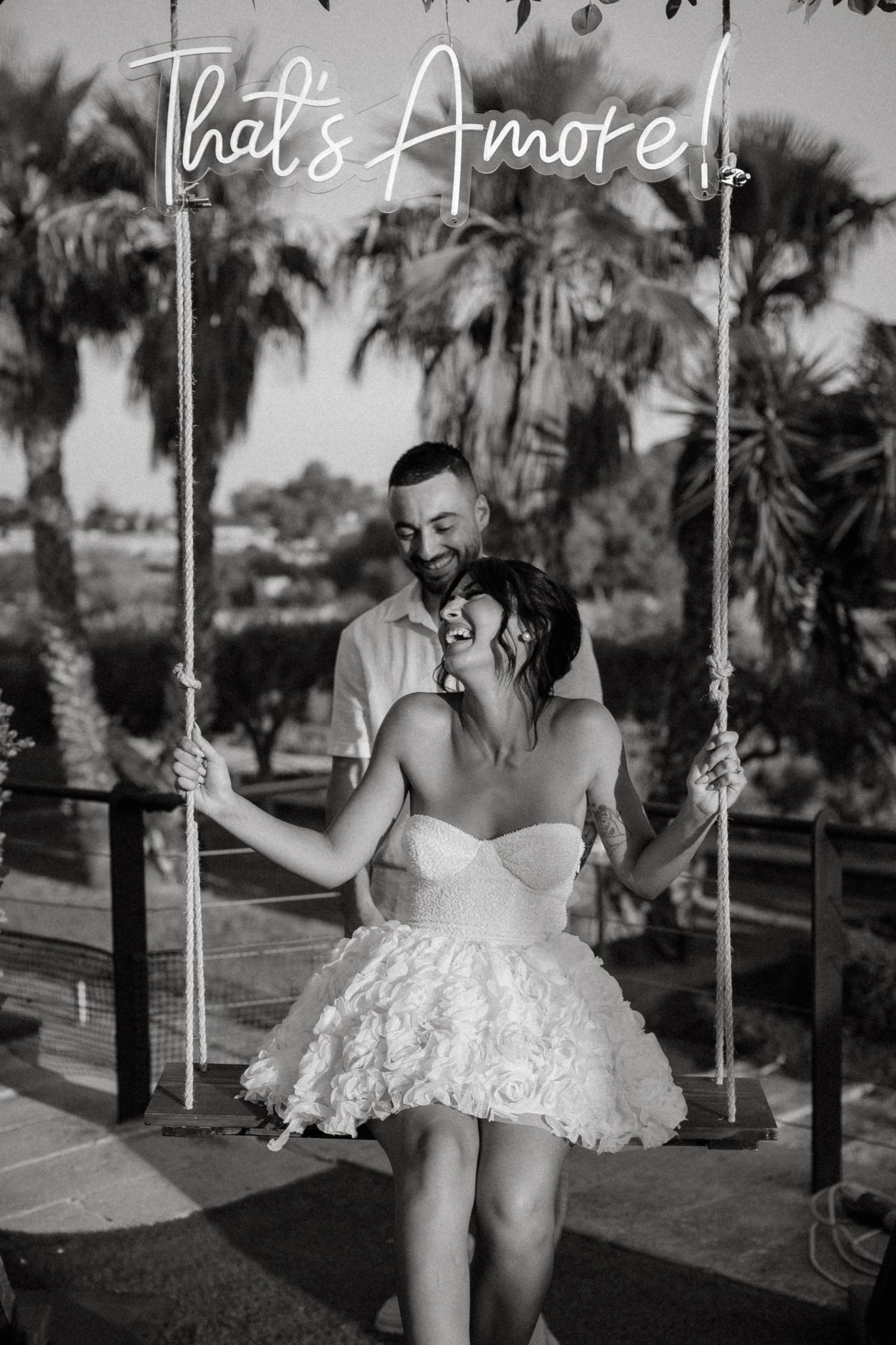 A playful black and white wedding photo of a couple on a swing under a neon sign.