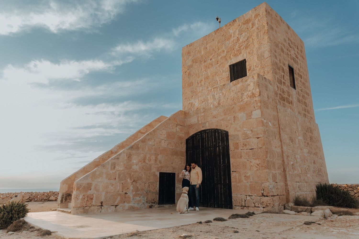 A rustic stone watchtower stands against a bright blue desert sky with scattered clouds.