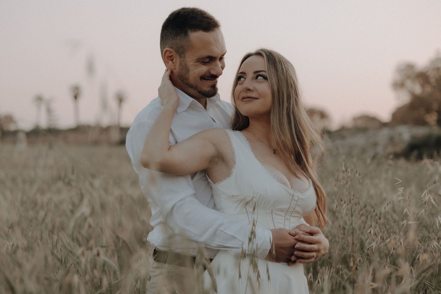 Romantic couple hugging in a wheat field at dusk, woman in white dress smiling up at man behind her.