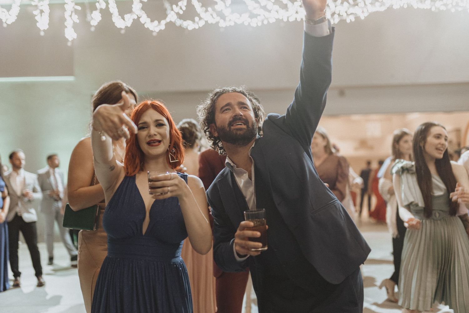 String lights sparkle overhead as guests celebrate at an indoor venue.