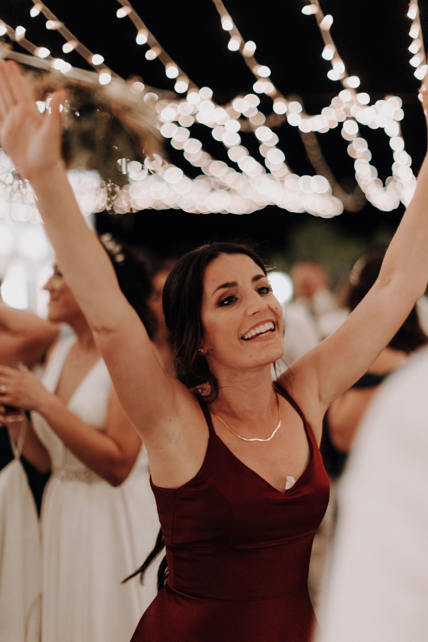 A woman in a burgundy dress dances joyfully under string lights at a wedding celebration.