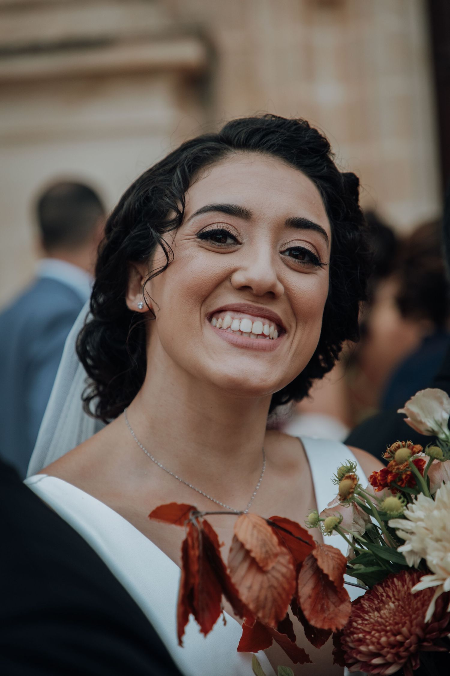 A radiant bride smiles while holding an autumn-toned bouquet featuring burgundy leaves and flowers.