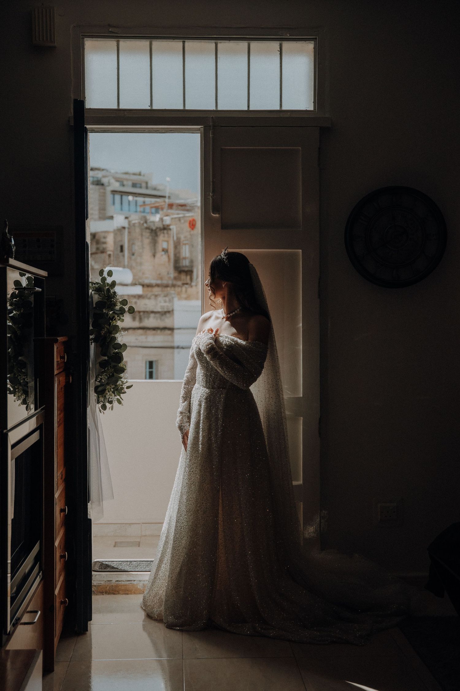 Silhouette of bride in wedding dress looking through window in moody lighting.
