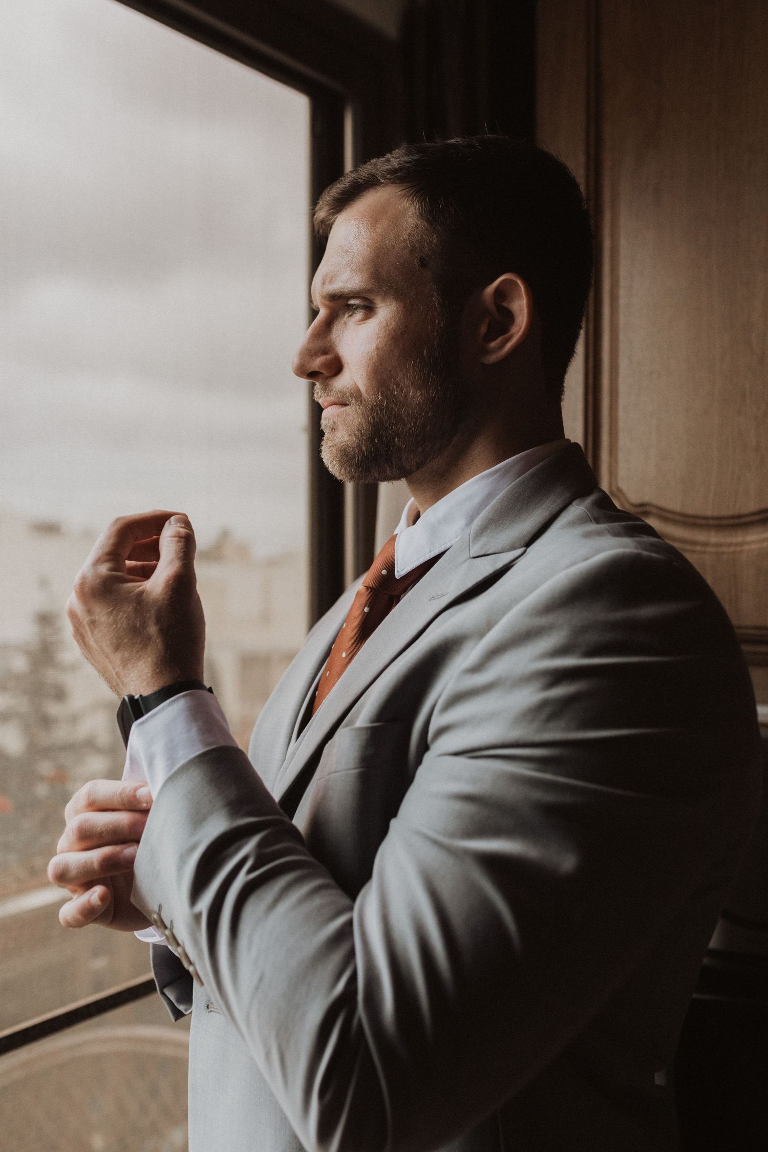 Person in grey suit looks thoughtfully out window during wedding preparations.