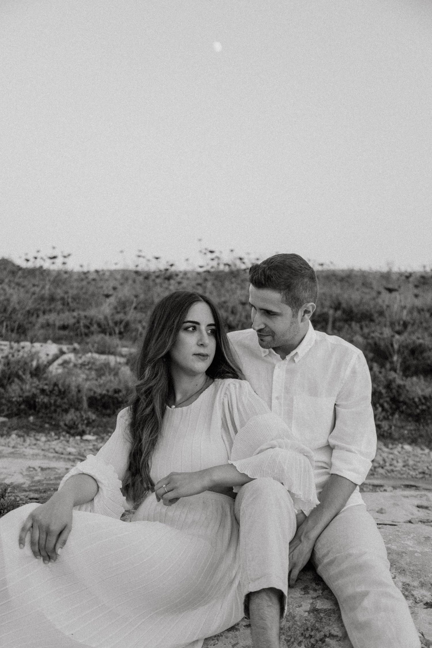 Black and white photo of a couple in white clothing sitting on rocky coastal terrain.