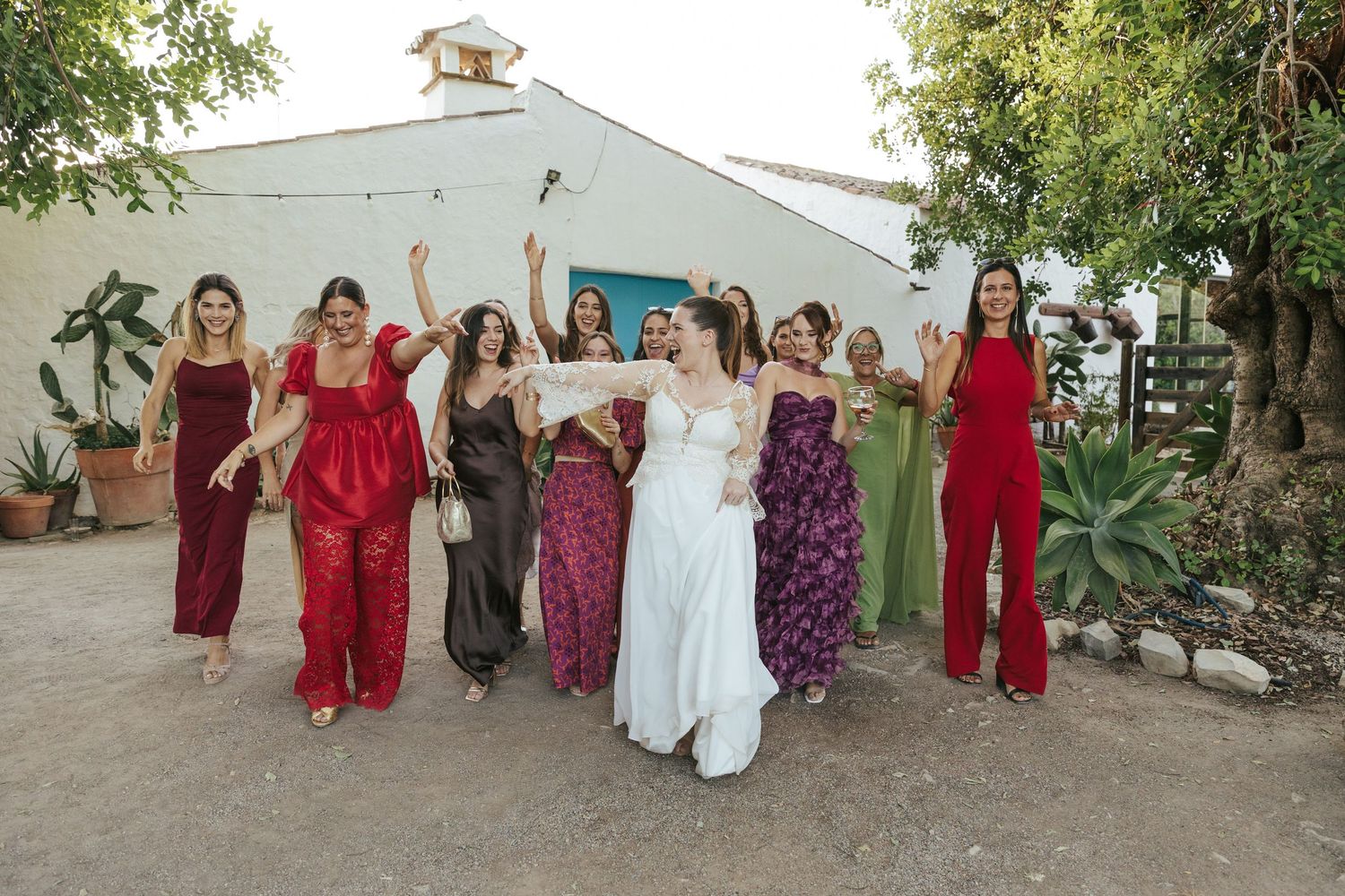 A group of people wearing colorful formal dresses and gowns pose together outdoors against a white wall.