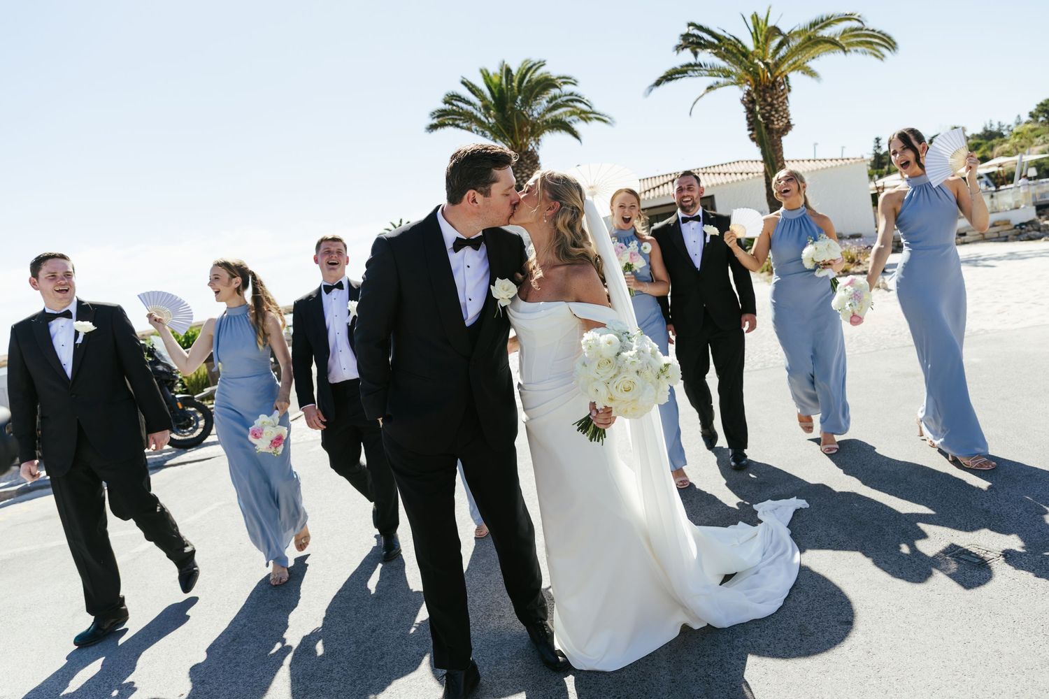 Bride and groom kissing surrounded by wedding party in blue gowns and black tuxedos with palm trees backdrop.