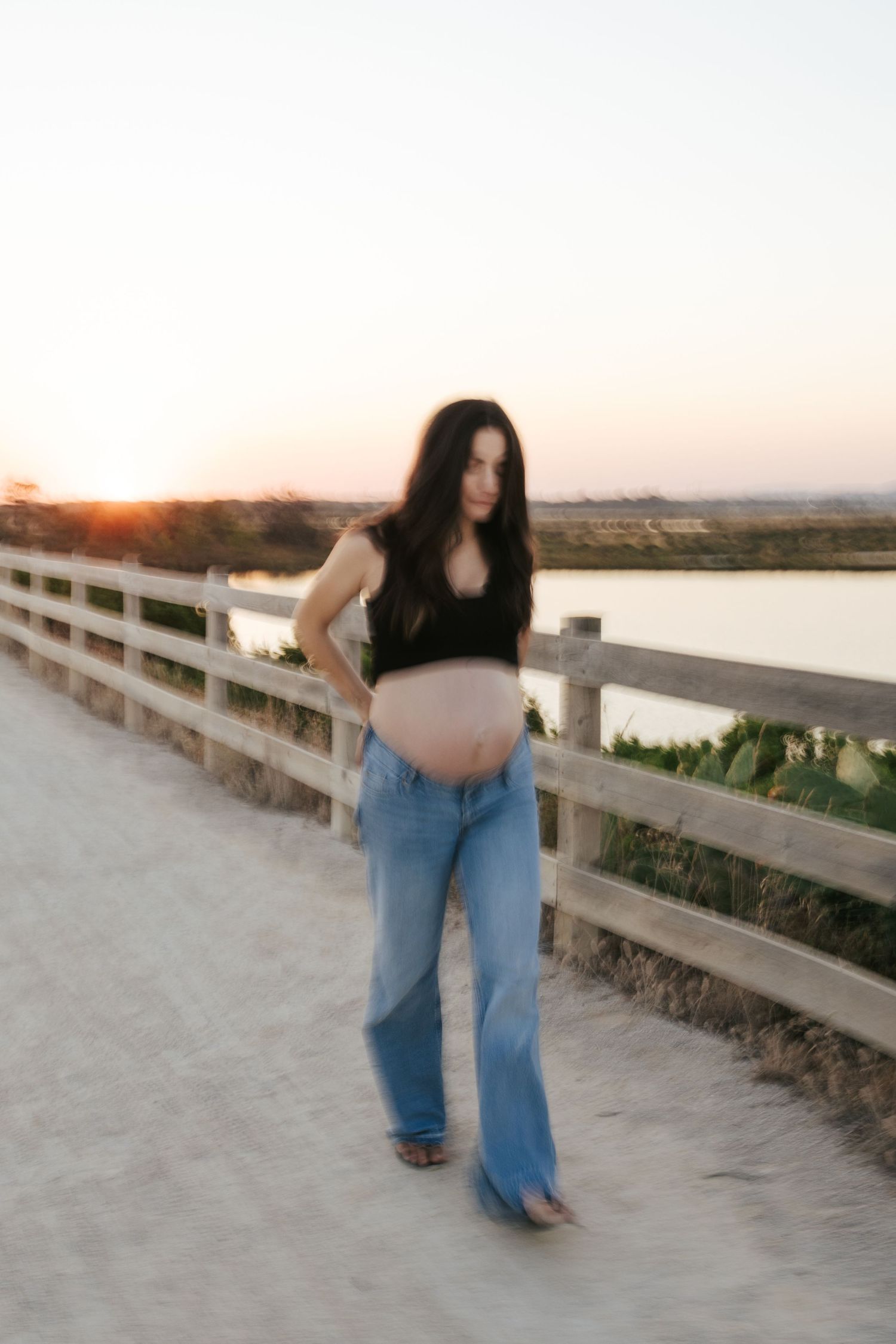 Pregnant woman in black crop top and blue jeans holds baby bump on sandy boardwalk path at sunset.