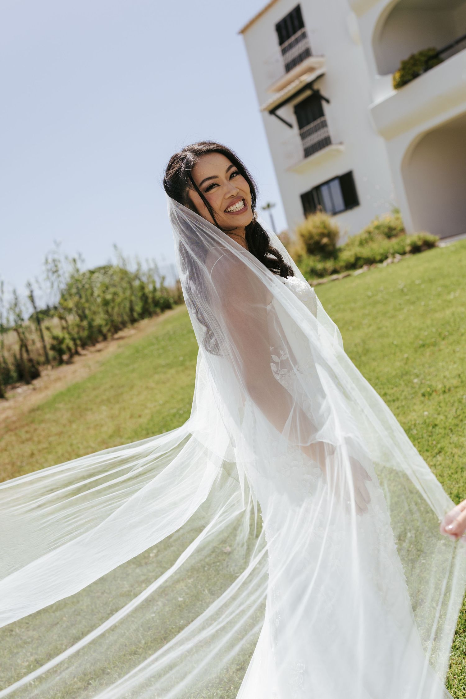 Smiling bride in white wedding dress and flowing veil twirling outdoors on green lawn near white building.