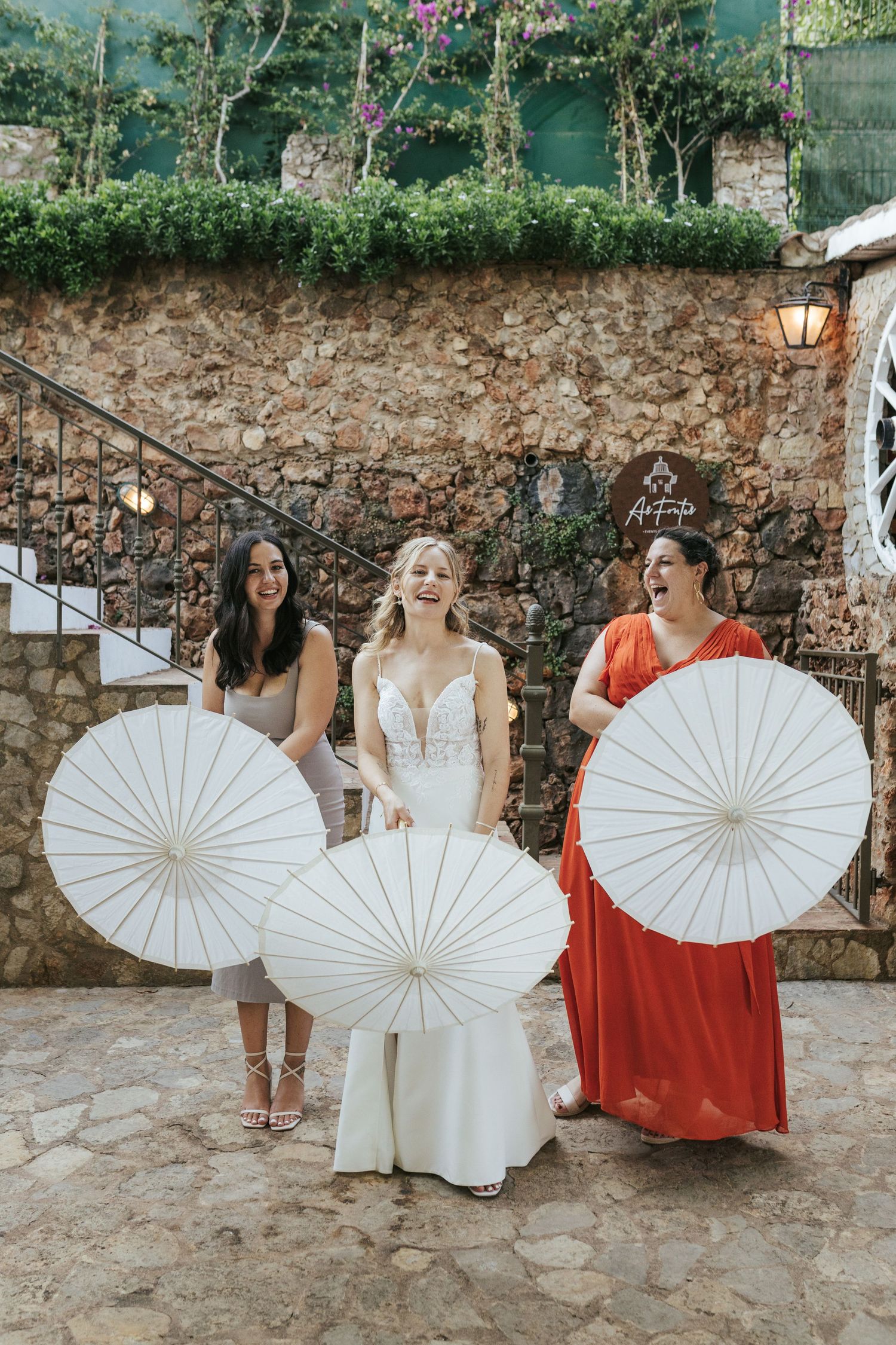 Wedding party holding white parasols poses against rustic stone wall with climbing flowers.