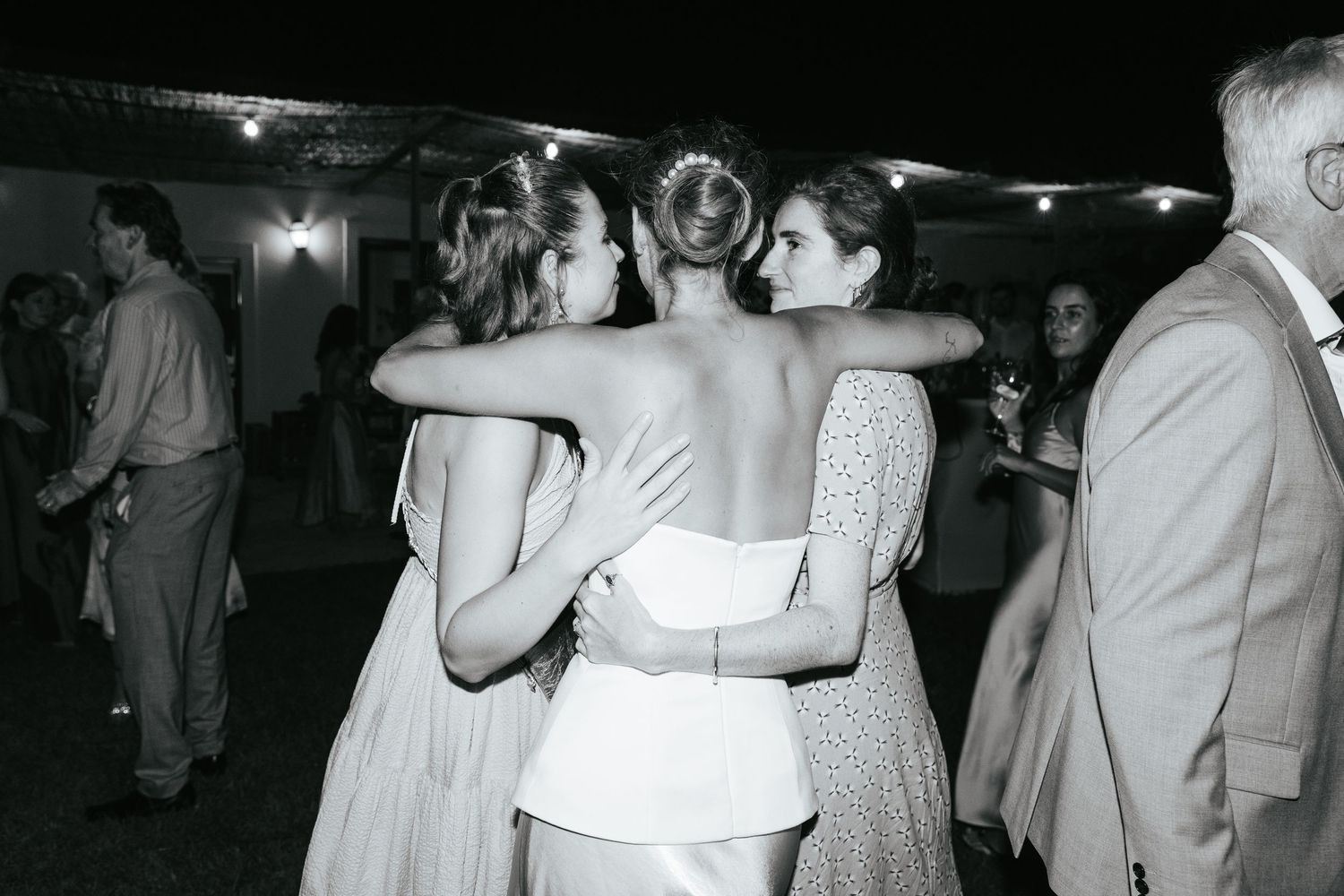 Bride embraced by two female guests in heartfelt hug at outdoor wedding reception in black and white.