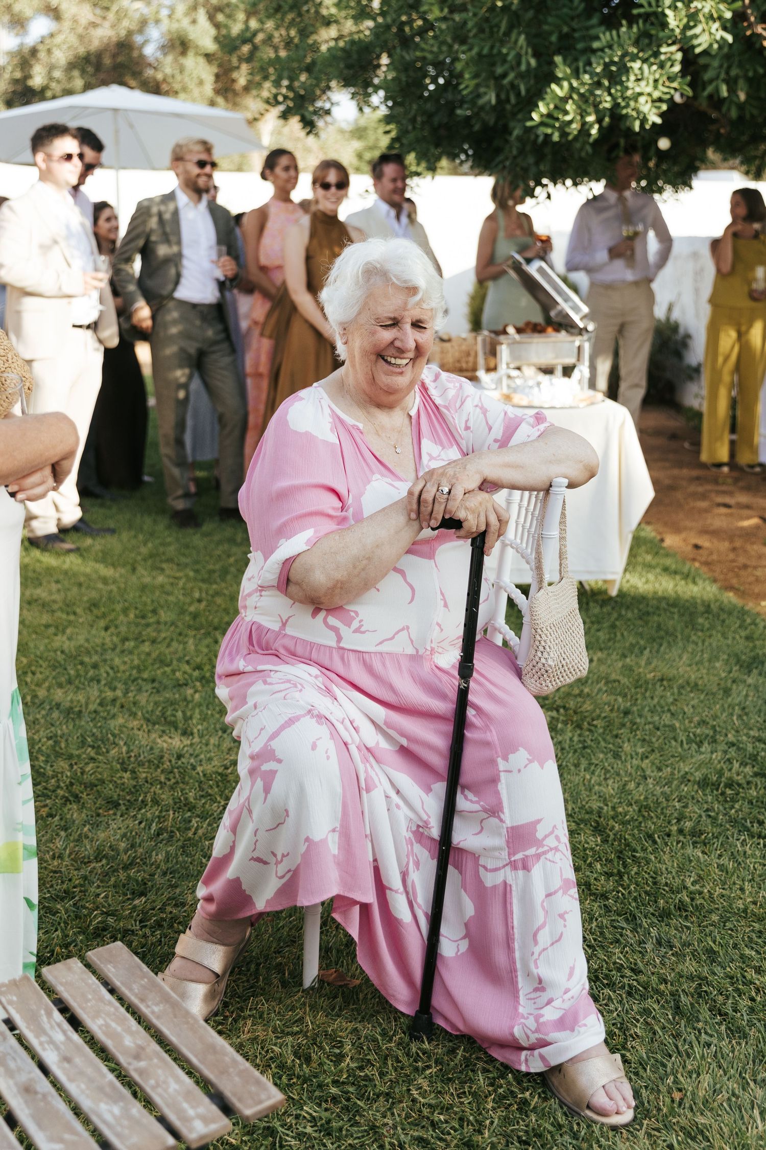 Elderly woman in pink floral outfit sitting with cane, laughing at outdoor wedding reception.