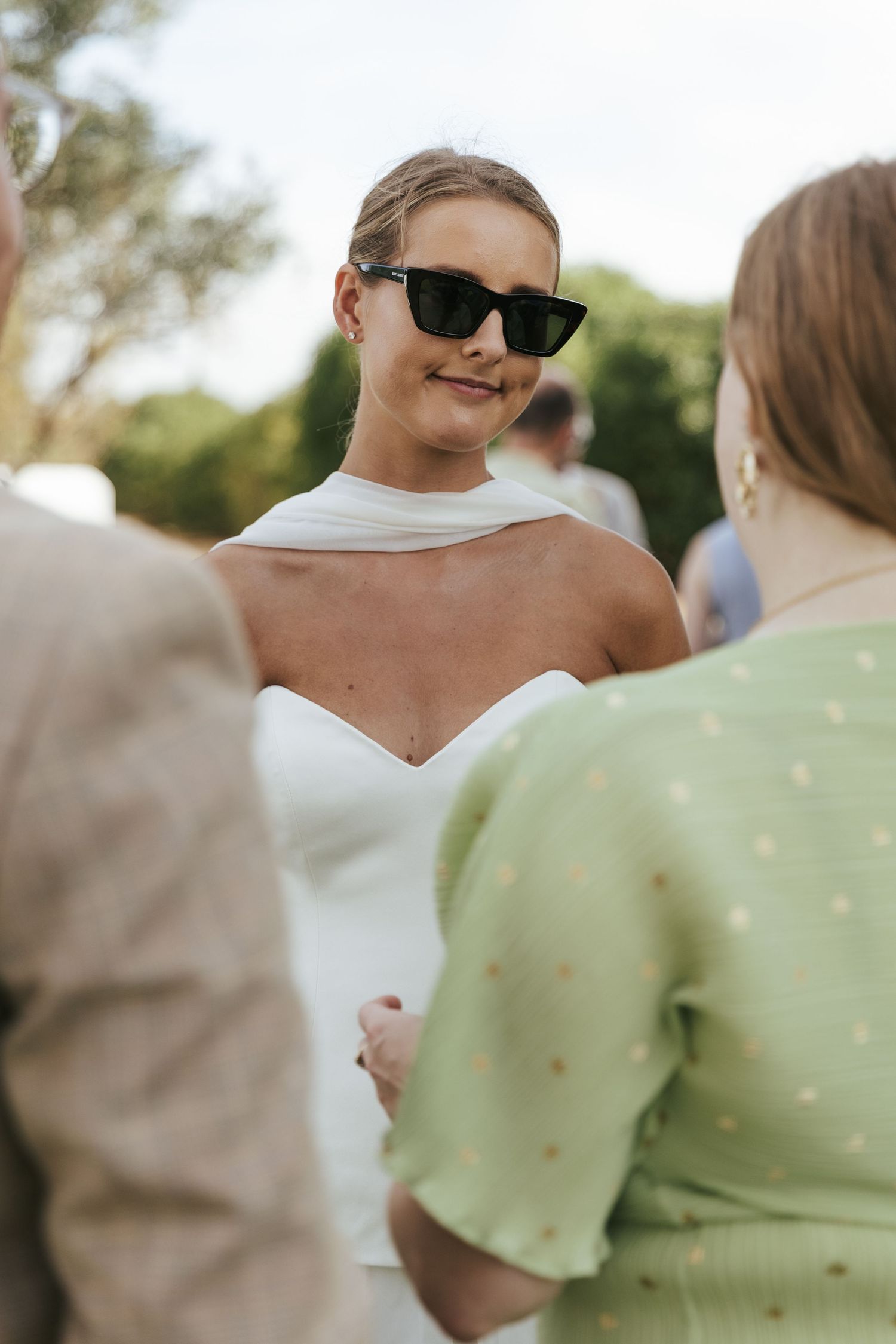Bride in white halter gown and black sunglasses smiling while chatting with guests at an outdoor wedding reception.
