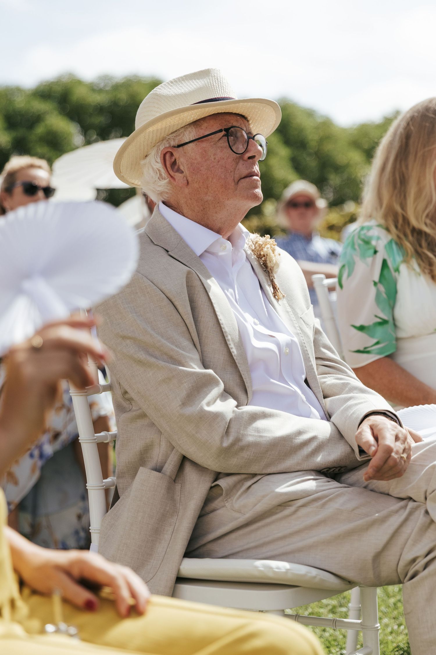 Elderly man in beige suit and white panama hat wearing glasses seated outdoors at a summer garden event.