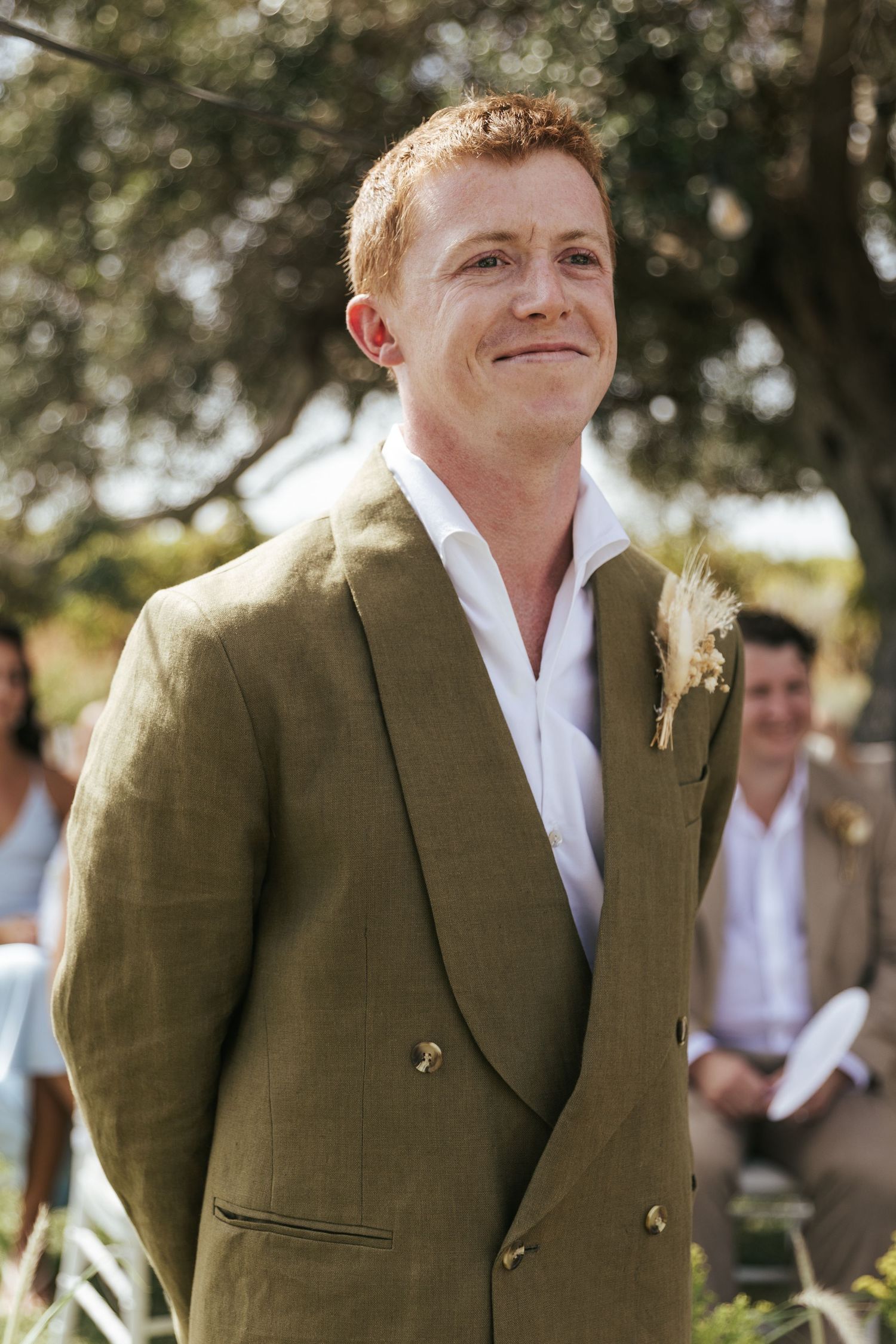 Groom in olive green double-breasted suit with boutonniere smiling emotionally at outdoor wedding ceremony.