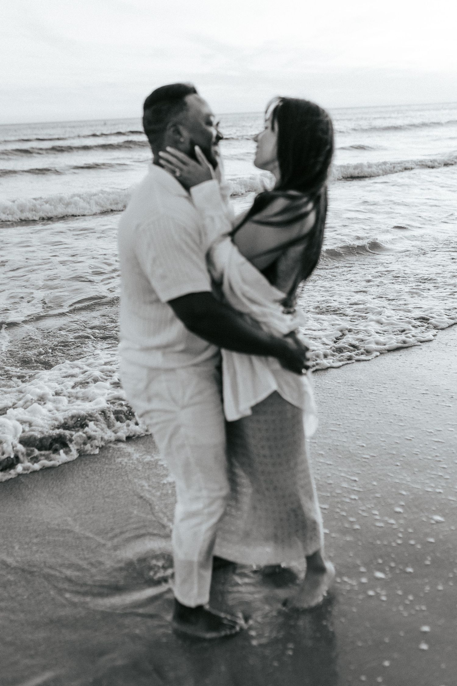 A couple in light colored clothing embraces romantically on the beach with ocean waves in the background.