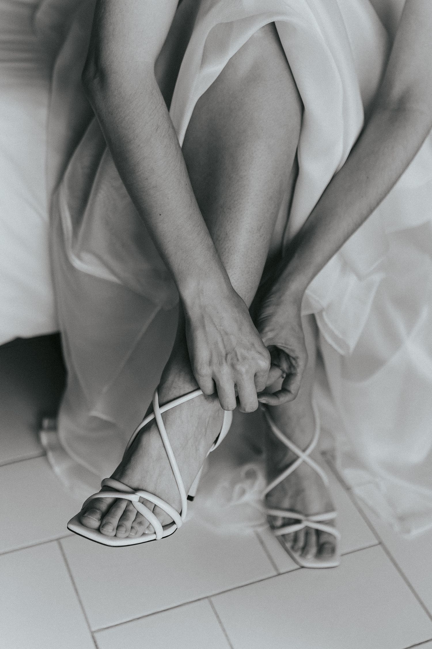 A black and white close-up shot of hands fastening elegant white strappy wedding shoes.