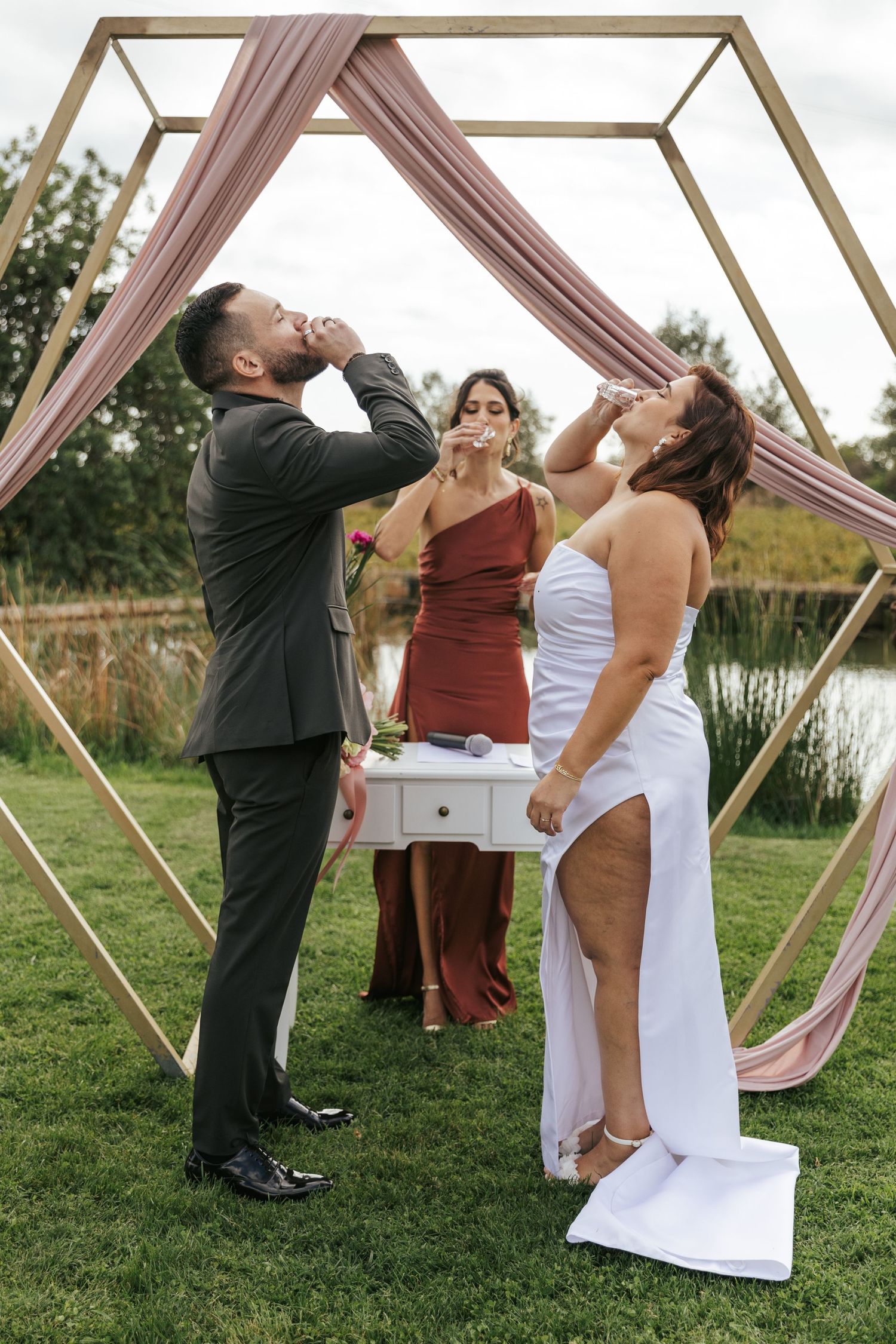 Wedding party members taking celebratory shots under geometric arch with pink draping by a lake.