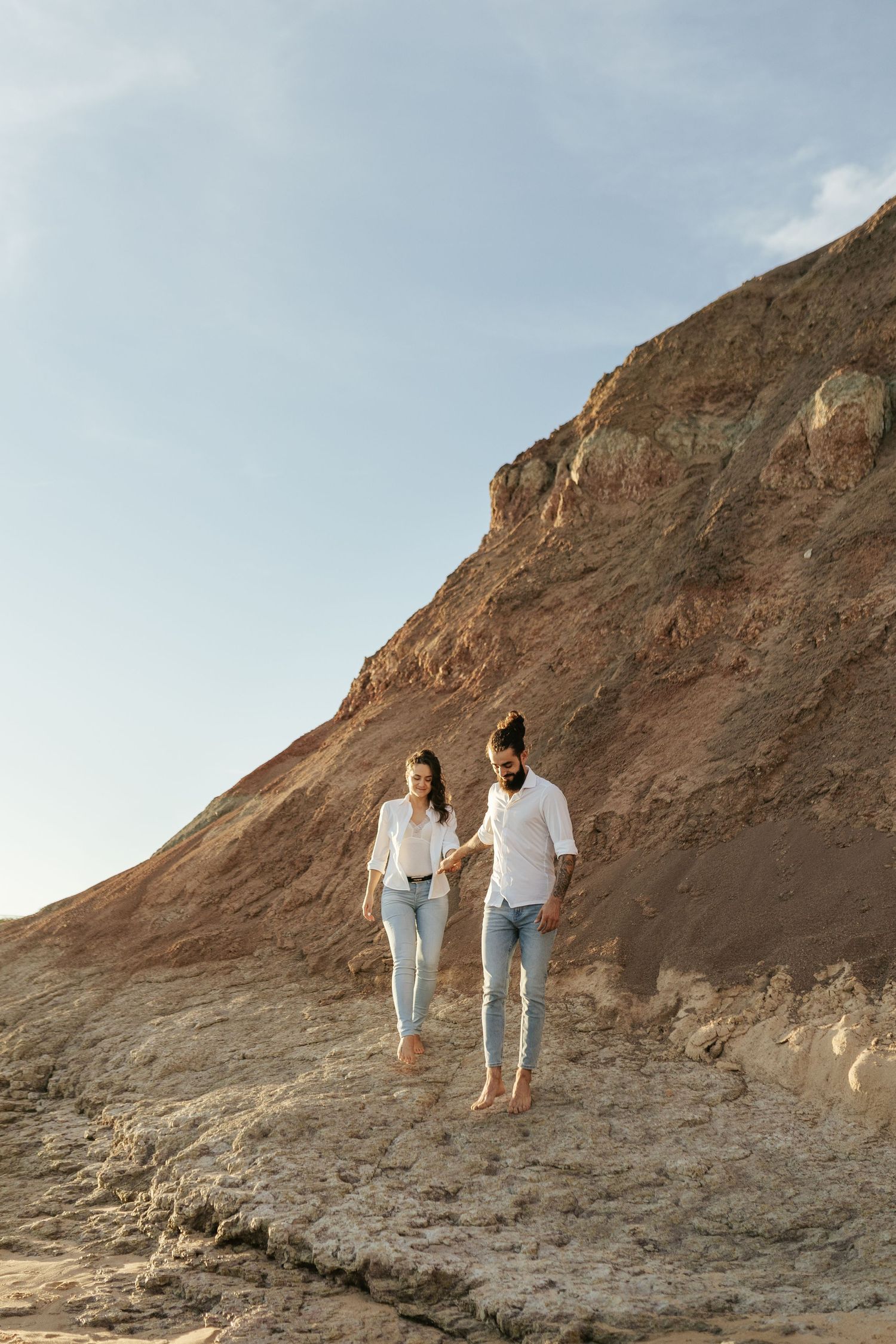 Couple walking barefoot along rocky cliff path during golden hour. Praia da Luz, Lagos, Algarve, Portugal.