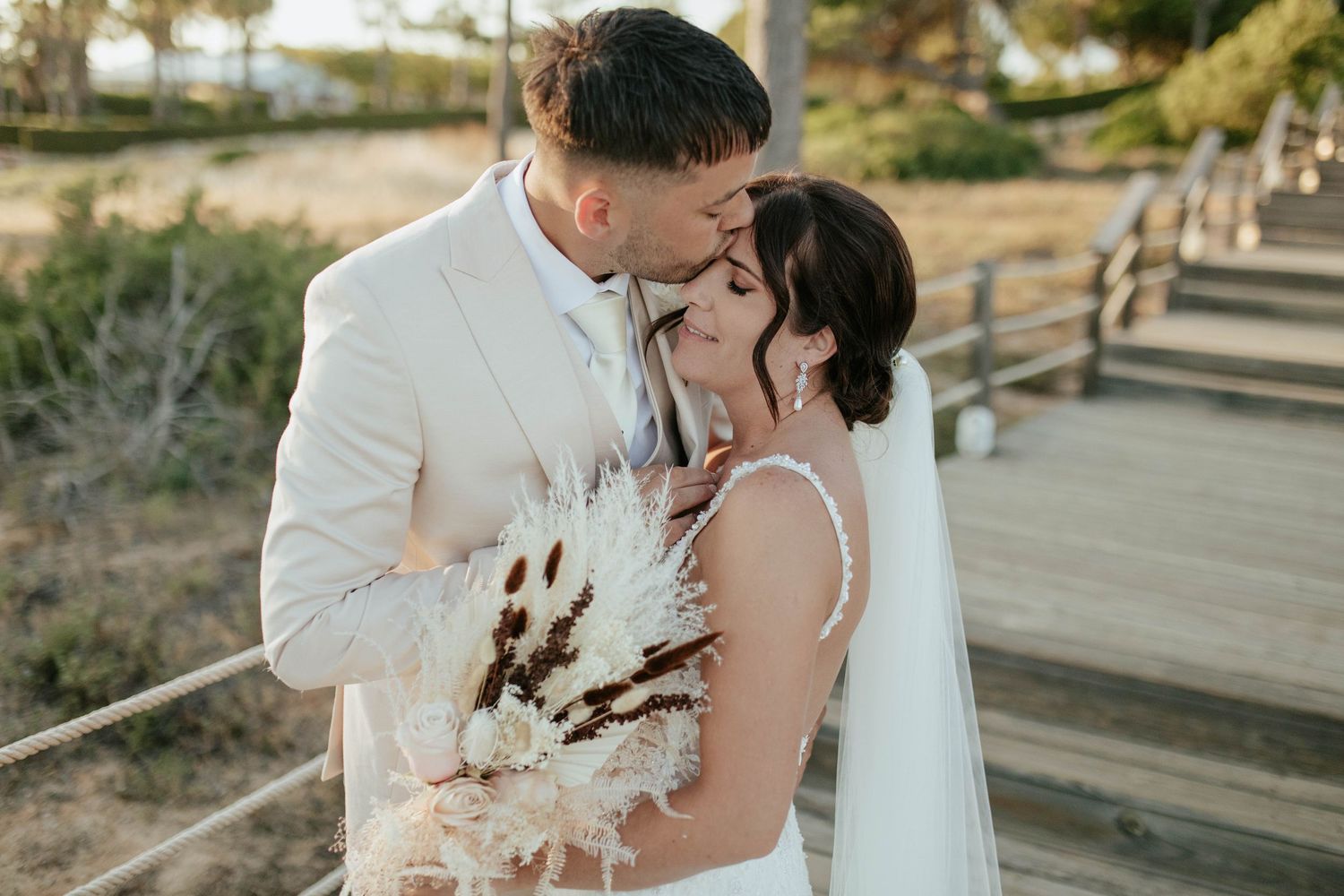 A couple in wedding attire shares a tender moment on a wooden walkway, with dried pampas grass bouquet at sunset.