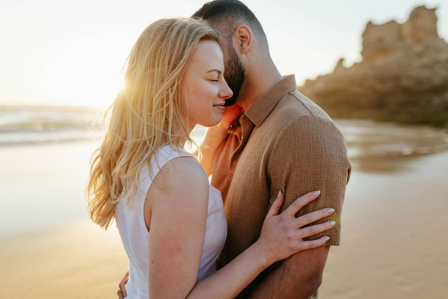 An intimate moment shared between a couple on a beach at sunset with rocky cliffs in the background.