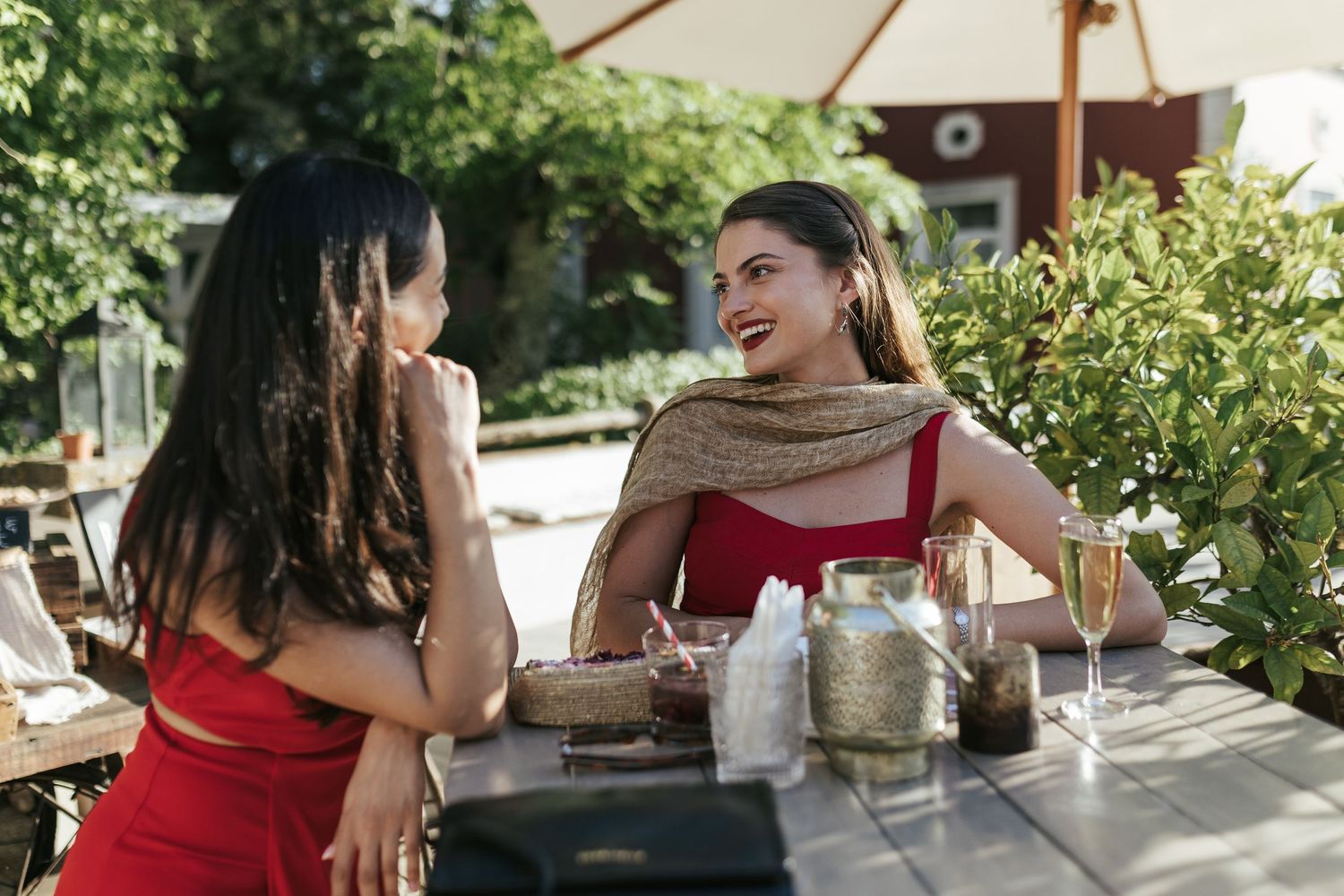 Two friends in red and beige attire having drinks and conversation at an outdoor cafe patio table on a sunny day.