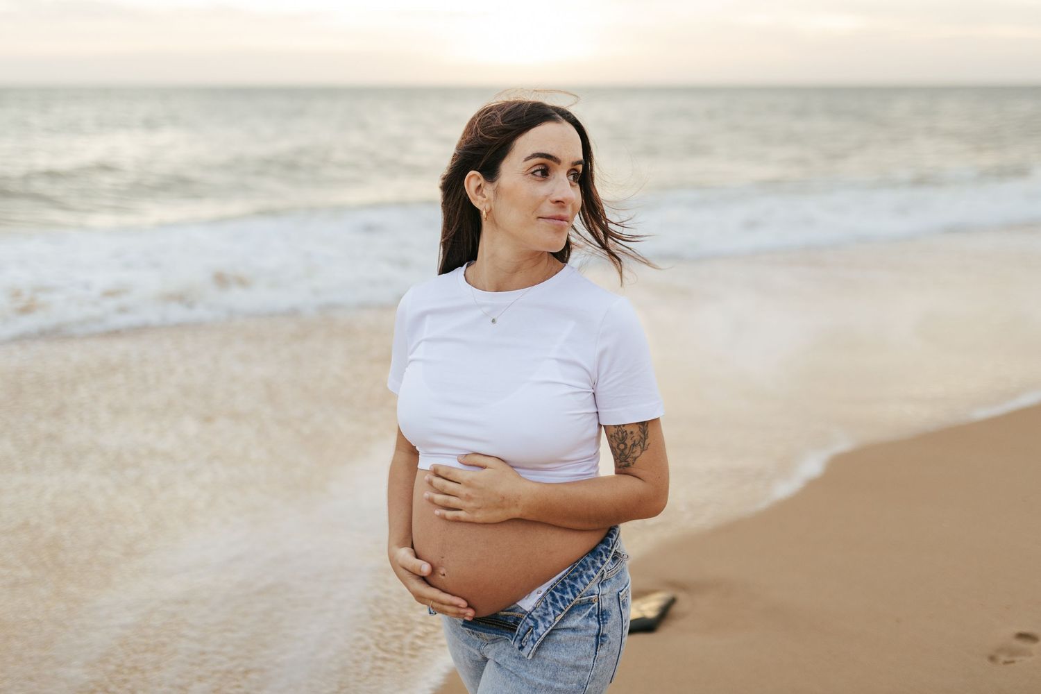 Someone wearing a white crop top and denim shorts stands thoughtfully on a sandy beach at sunset.