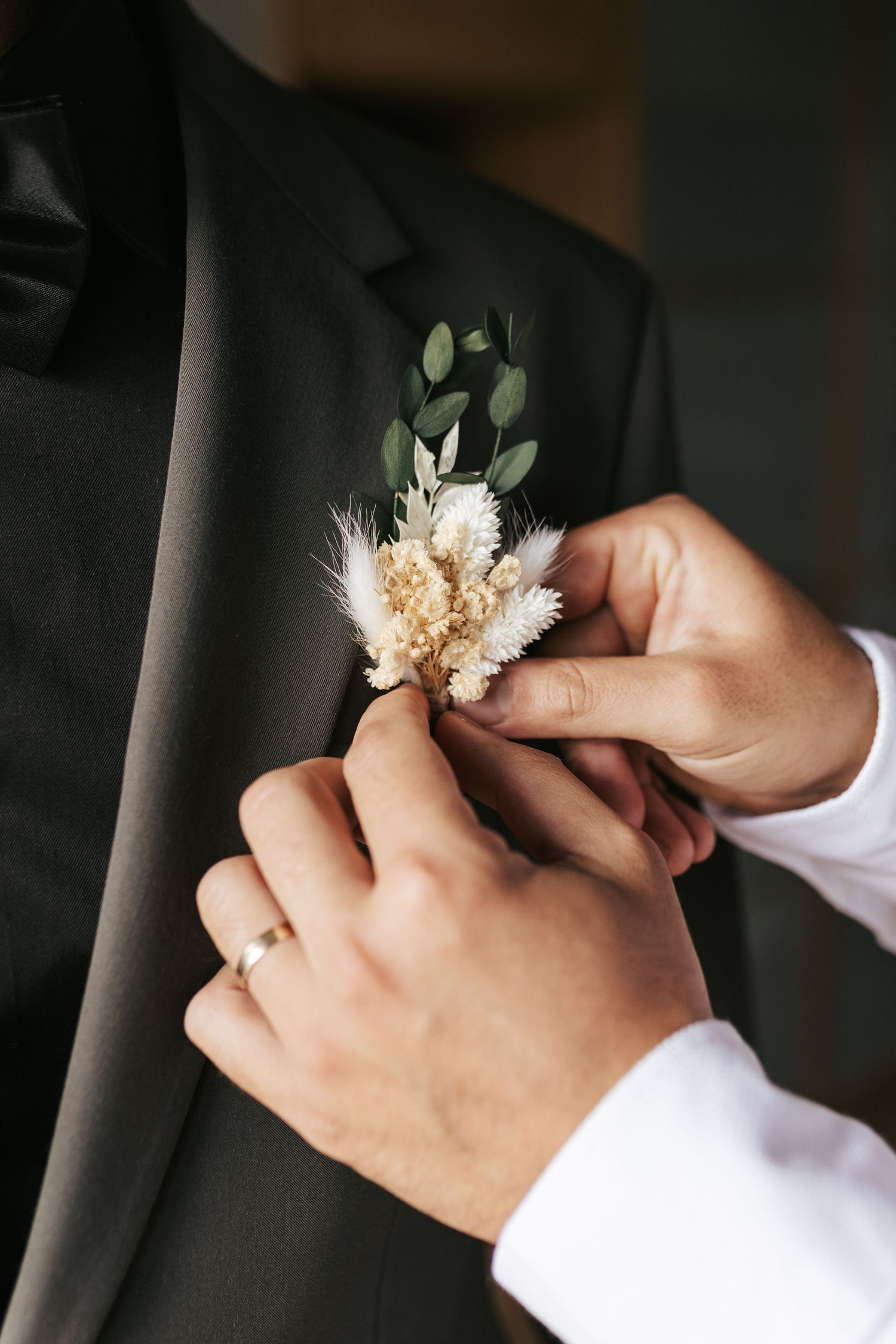 Hands pinning a delicate dried flower and greenery boutonniere to a black suit lapel.