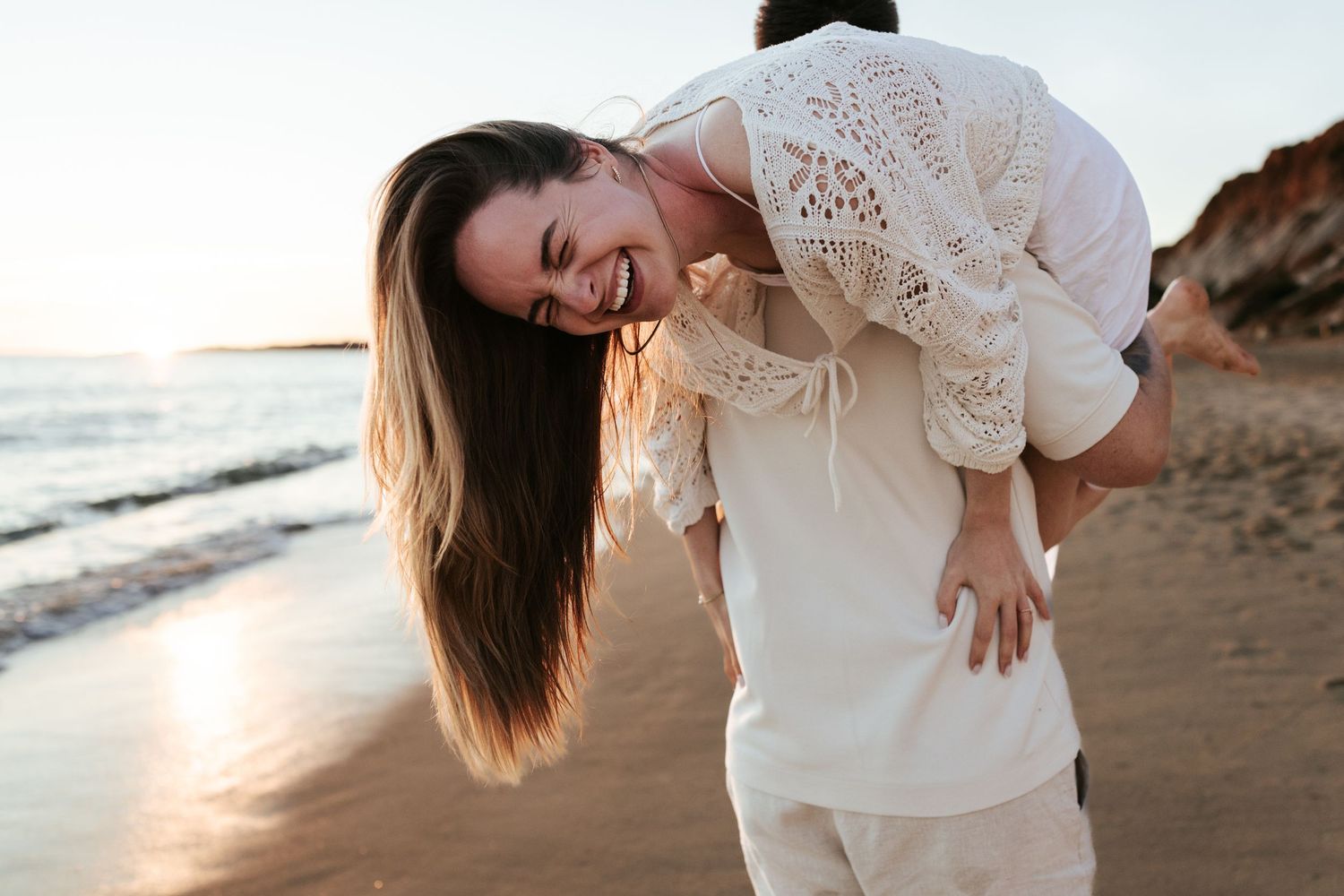 A joyful couple shares a romantic moment on the beach at sunset with waves and cliffs in the background.
