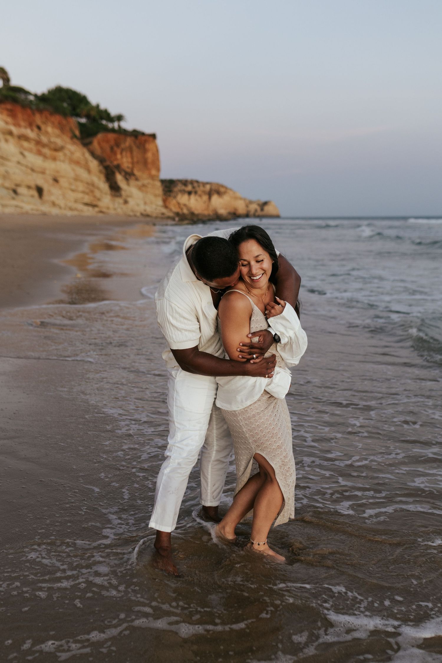 A couple shares an intimate embrace while standing in shallow ocean waves near coastal cliffs. Praia do Porto de Mós, Lagos, Algarve, Portug