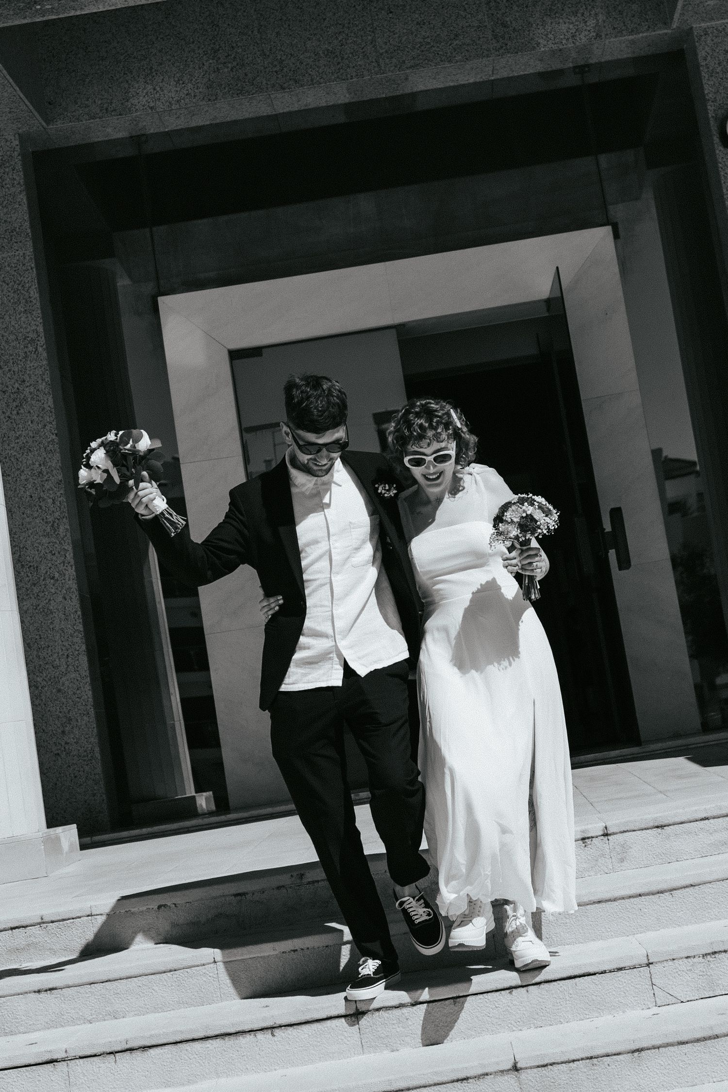 A joyful newlywed couple exits a building and jumps down steps in their wedding attire in black and white.