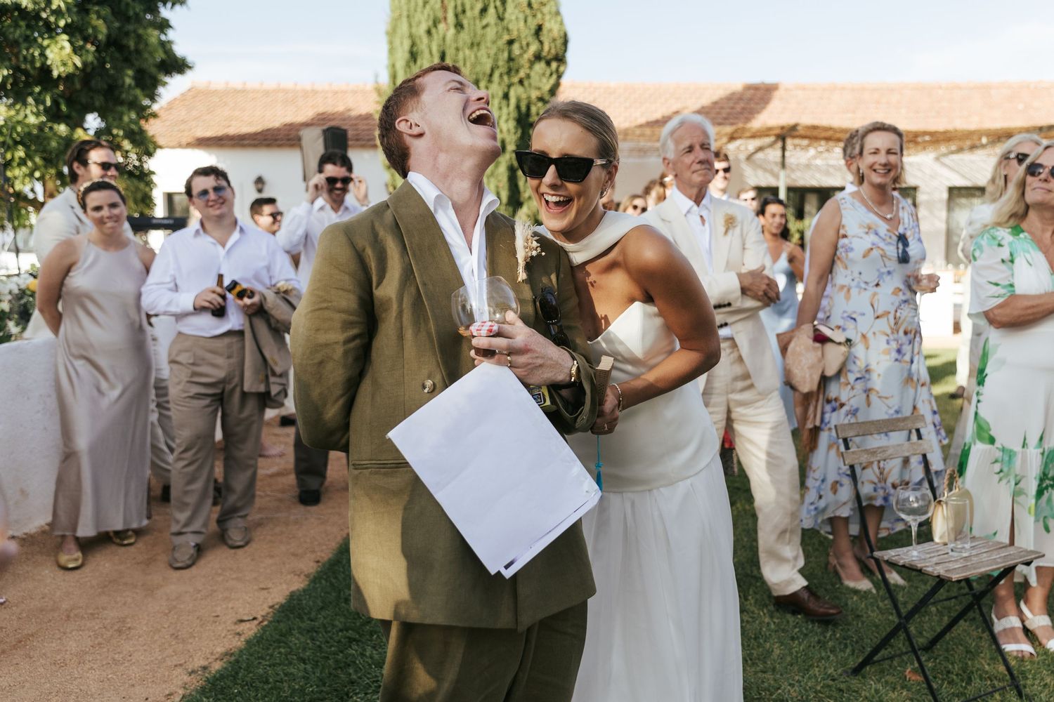 A group celebrates at an outdoor wedding reception with laughter and joy under green trees and sunlight.