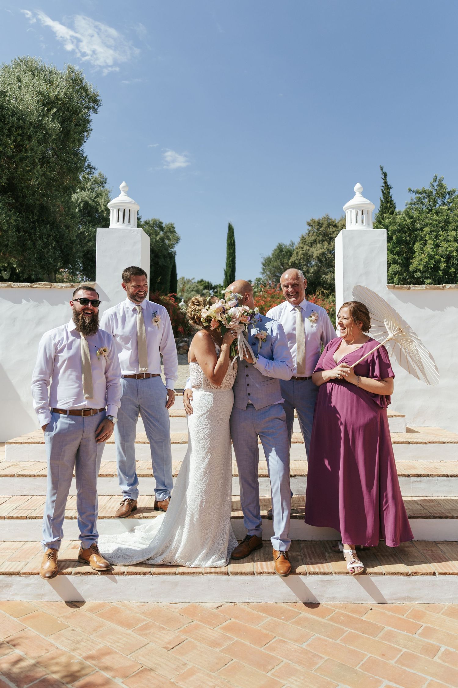 Wedding party poses on sunny steps, bride and groom kissing, groomsmen in lilac suits, woman in magenta gown holding parasol.