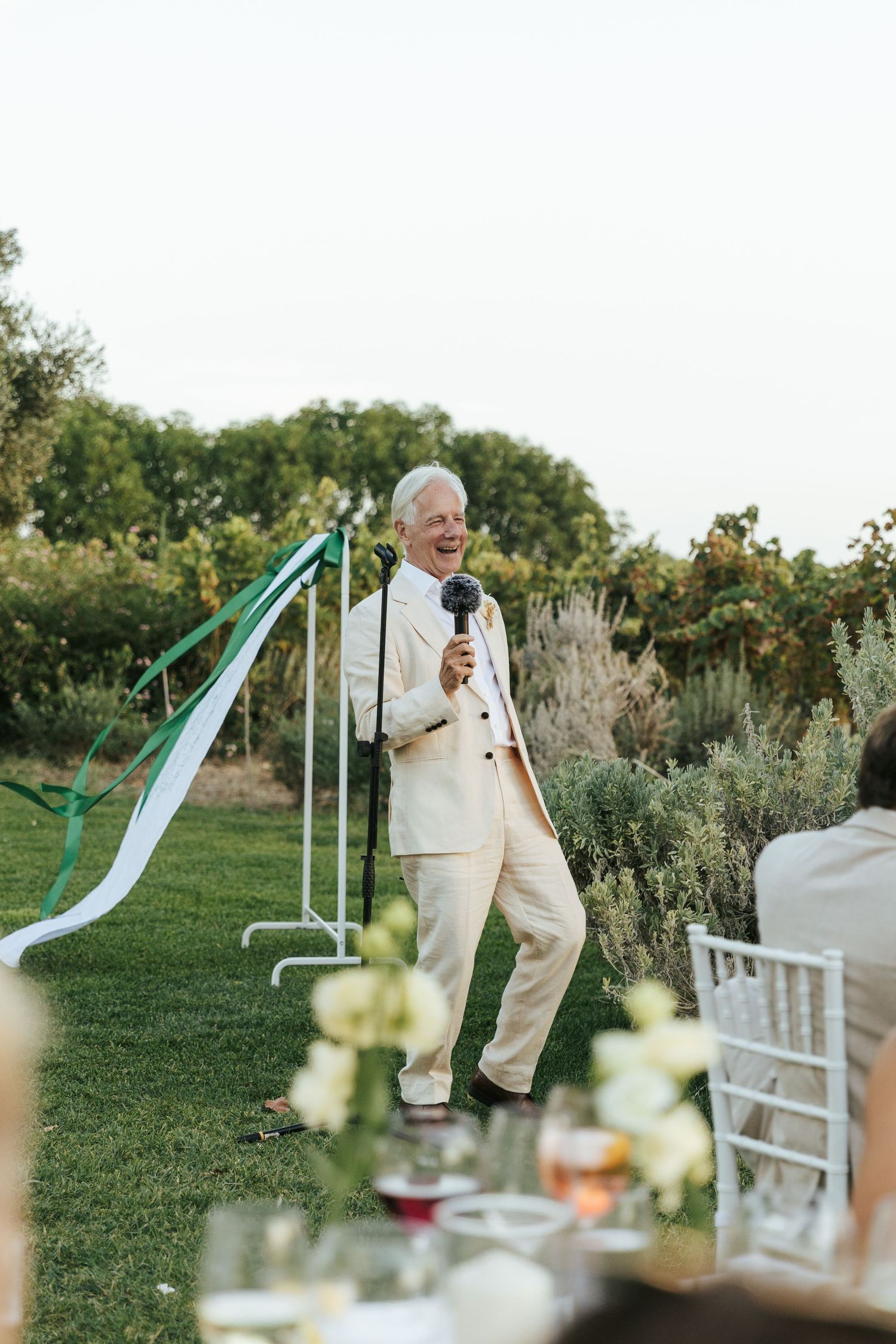 Elderly man in cream suit giving a speech at an outdoor wedding reception.