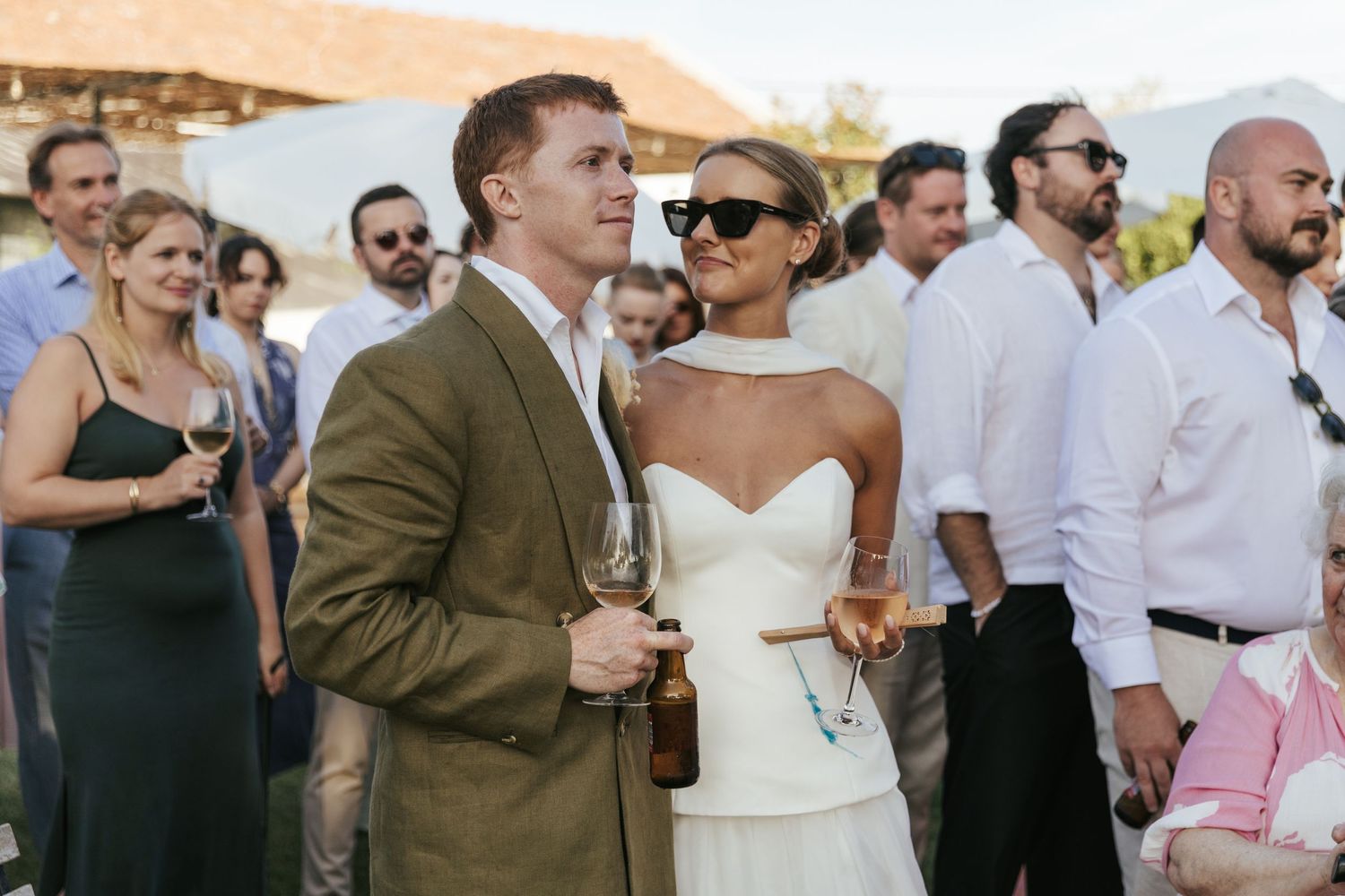 Bride in white strapless dress and groom in tan suit watching wedding reception outdoors.