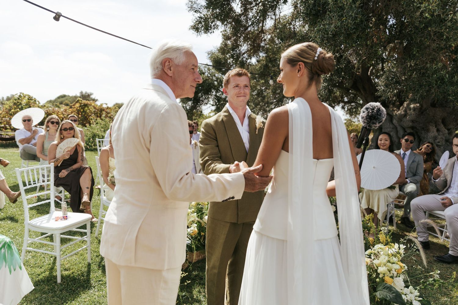 Bride and groom holding hands during outdoor wedding ceremony with guests seated on white chairs.