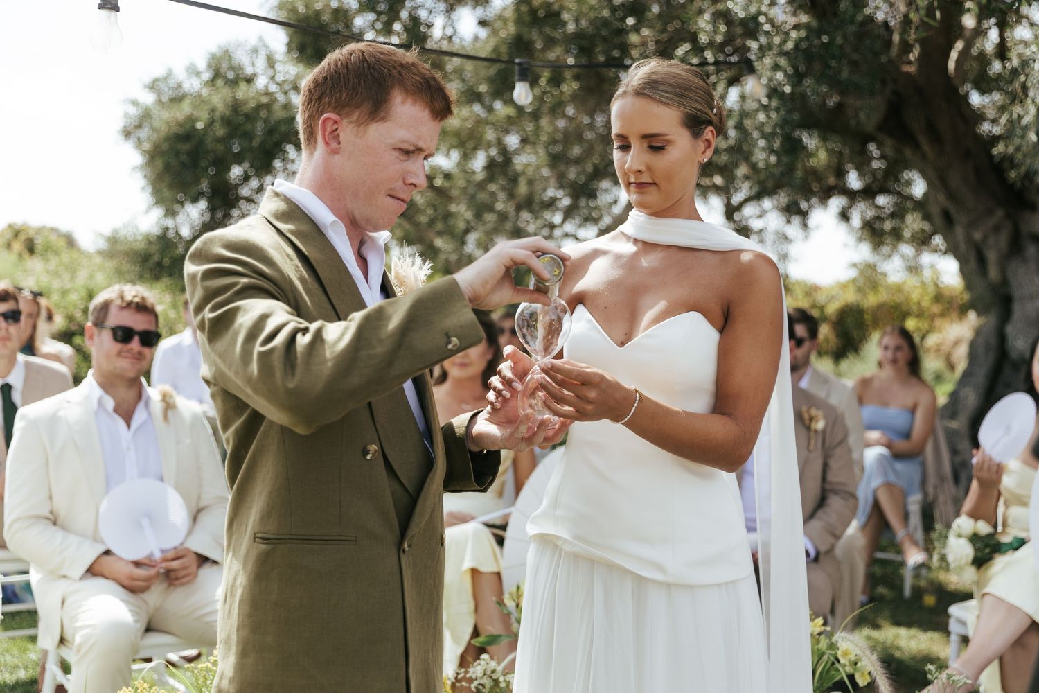 Groom in olive suit placing wedding ring on bride in strapless white gown during outdoor ceremony.