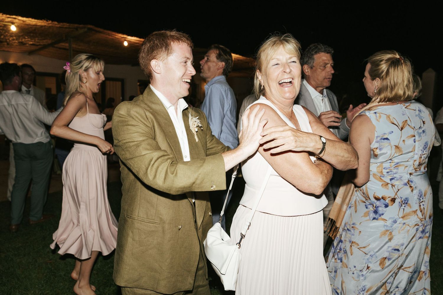 Groom in tan suit dancing and laughing with older woman at outdoor evening wedding reception.