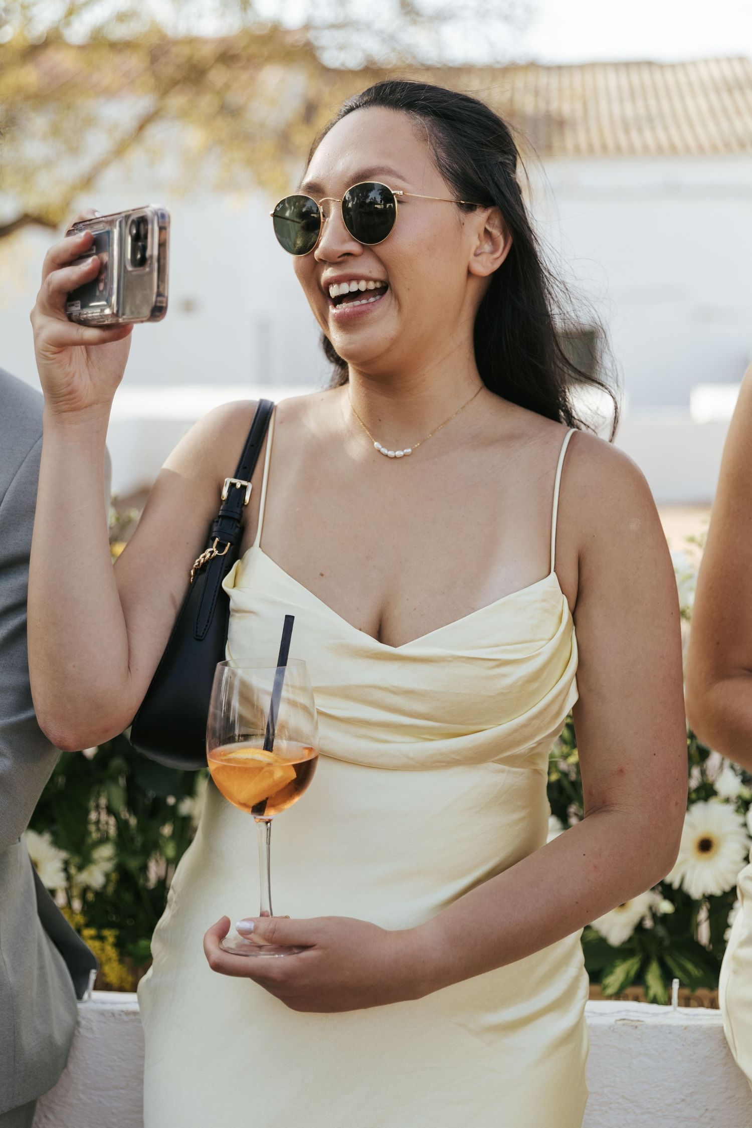 Smiling Asian woman in yellow slip dress holding wine glass and camera at a wedding reception.