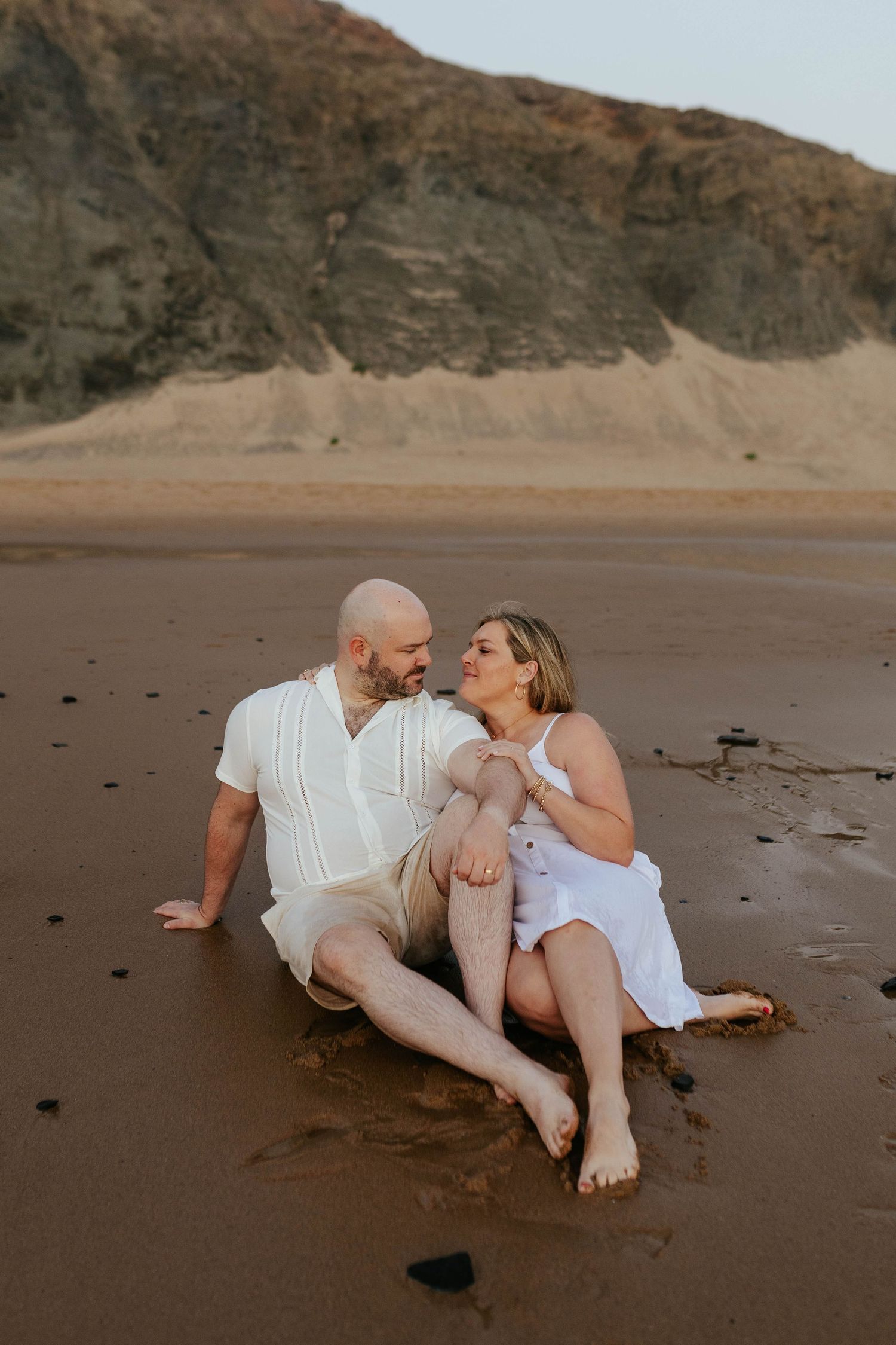 A couple sits together laughing and embracing on a sandy beach with dramatic cliffs in the background at sunset.