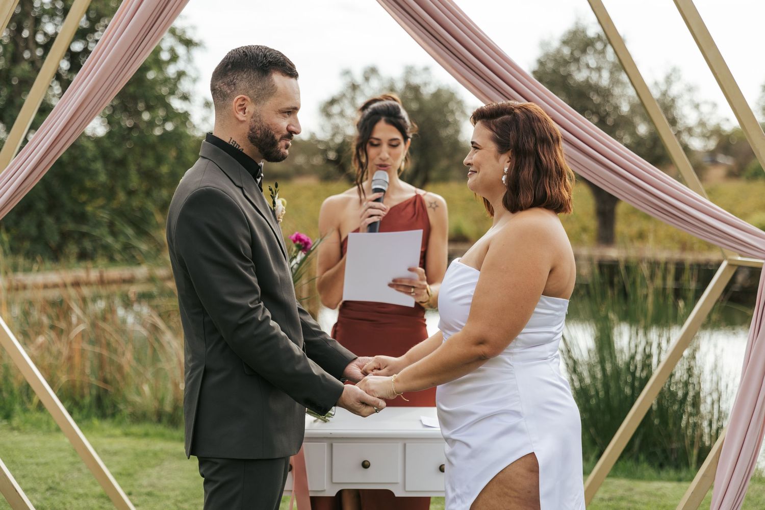 An outdoor wedding ceremony under a draped pink arch near a pond.