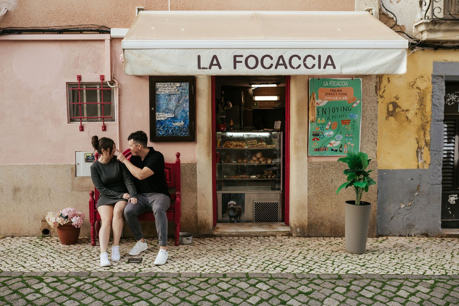 Two people sit on a bench outside La Focaccia shop storefront with pink walls and cobblestone streets in a European setting.