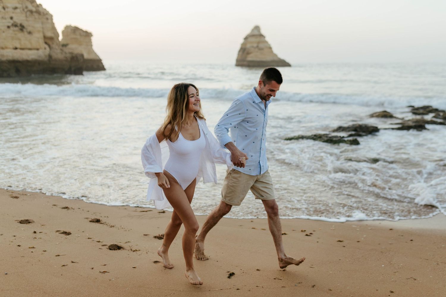 Joyful couple runs along sandy beach with dramatic coastal cliffs in the background. Praia Dona Ana, Lagos, Algarve, Portugal.