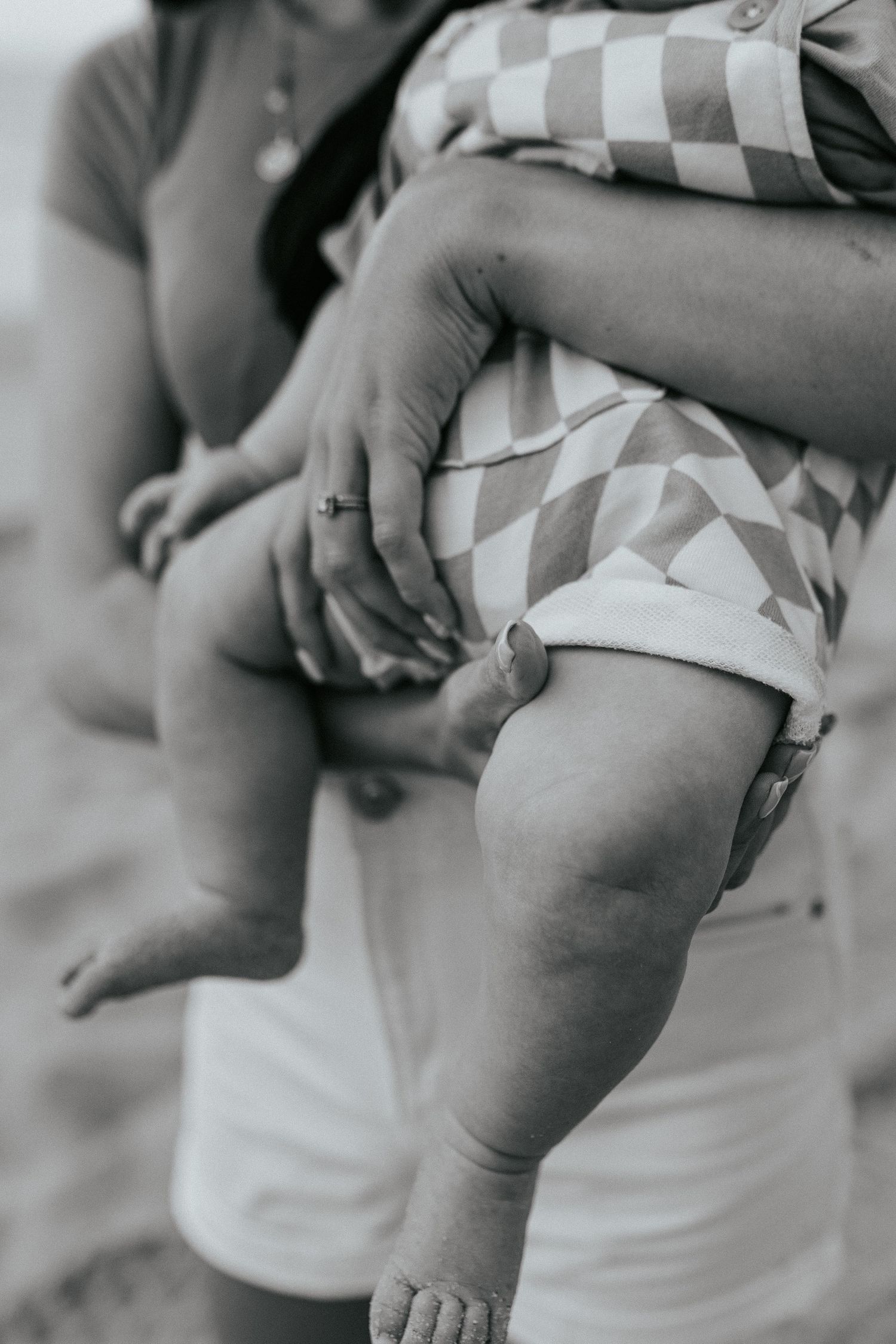 Close-up black and white image of a baby's chubby leg in checkered shorts with someone's hand tenderly touching it.