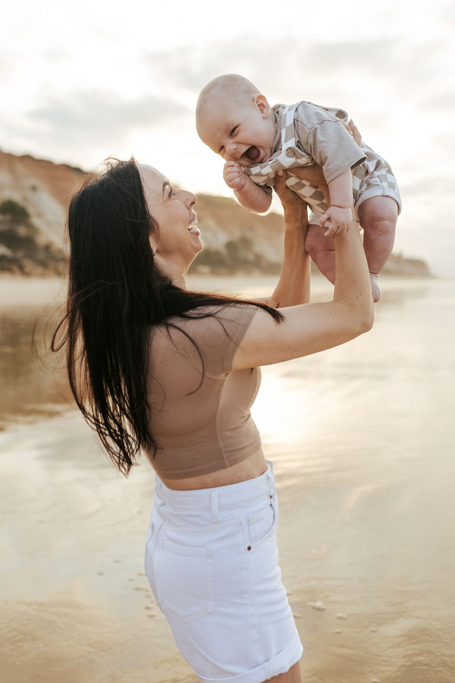 A loving parent lifts and plays with a laughing baby at sunset on a sandy beach with golden light.
