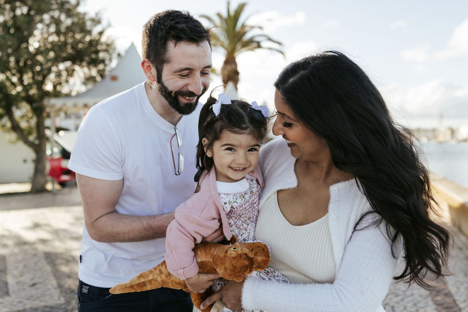 A couple and their toddler share a heartwarming moment together at a beach with palm trees in the background.