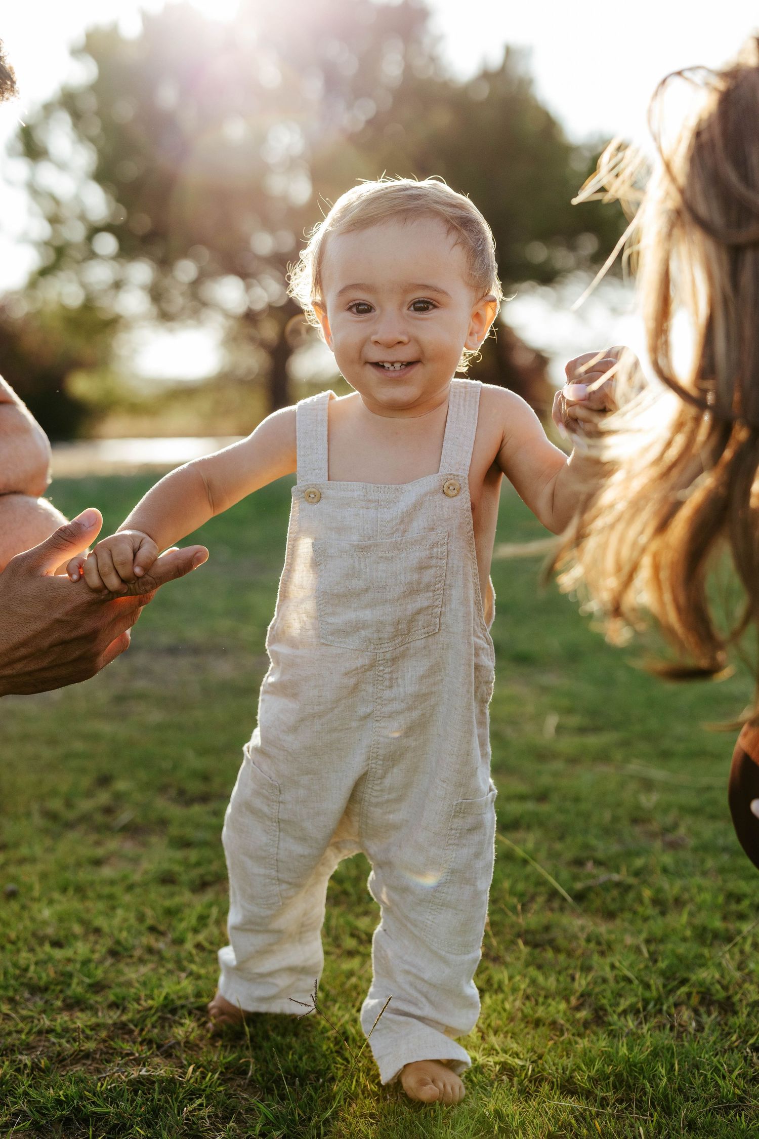 A small child in beige overalls holds hands with others while standing on grass in warm sunset lighting.