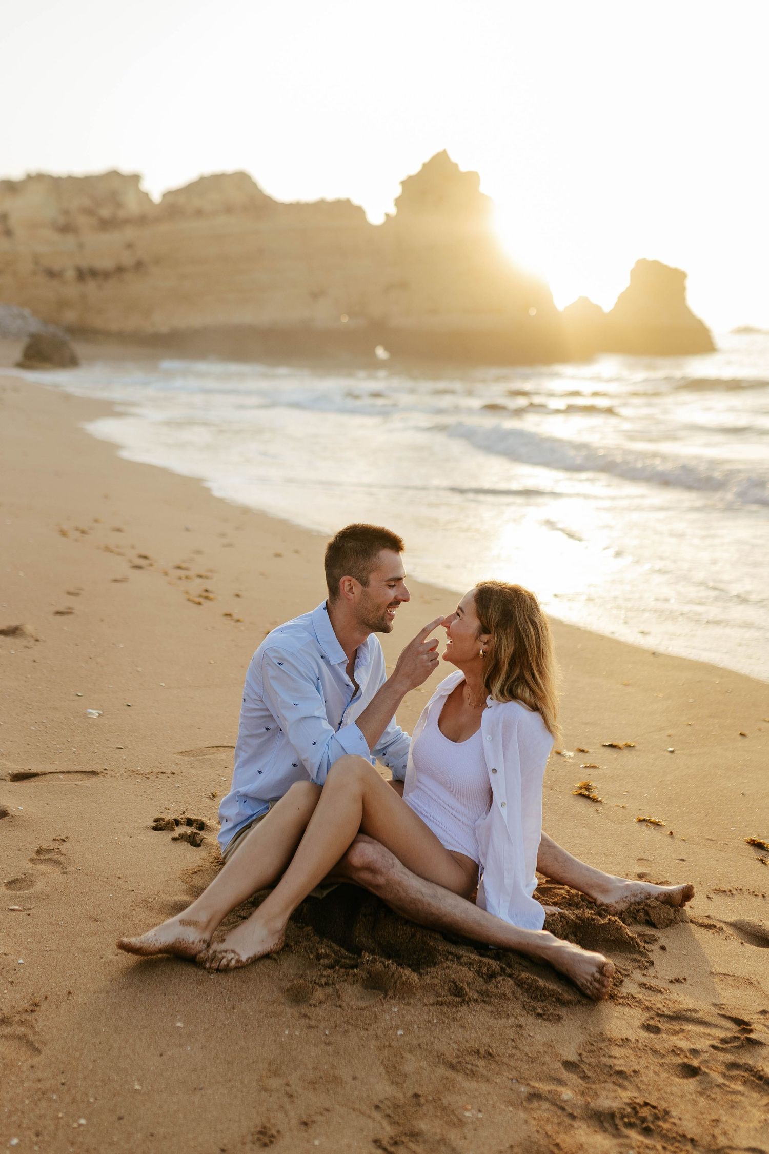 A couple laughs together while sitting on a sandy beach during sunrise. Praia Dona Ana, Lagos, Algarve, Portugal.