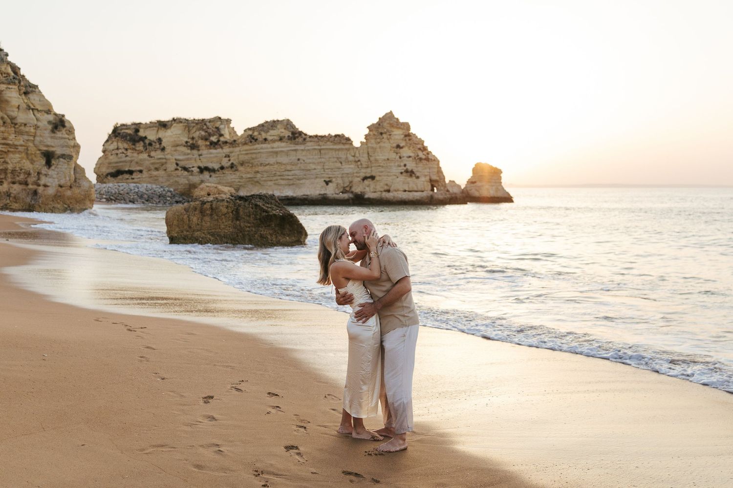 Couple embraces on sandy beach at sunset with dramatic limestone cliffs and ocean waves in Portugal.